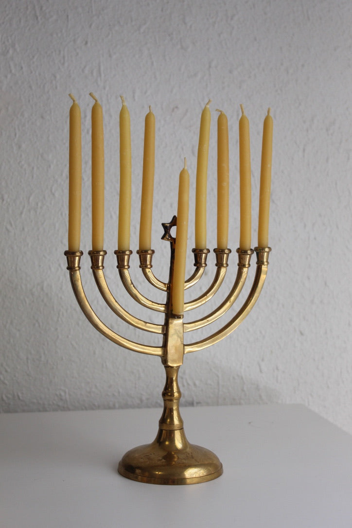 A brass Hanukkah menorah with nine lit yellow candles, displayed on a white surface against a textured white wall.