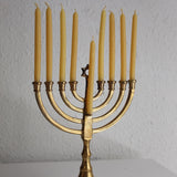 A brass Hanukkah menorah with nine lit yellow candles, displayed on a white surface against a textured white wall.