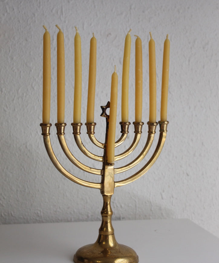 A brass Hanukkah menorah with nine lit yellow candles, displayed on a white surface against a textured white wall.